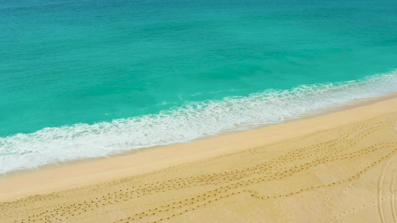 Aerial view of the Dreams Beach shoreline where the white surf band sits close to the pale beach, with patterned footprints on the dry sand and no visible people.