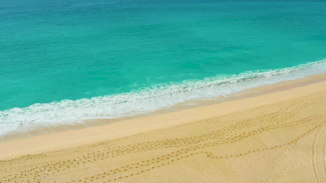 Aerial frame from Dreams Beach in Baja California Sur with blue water across the top, a diagonal white wave edge, pale sand below, and no visible people.