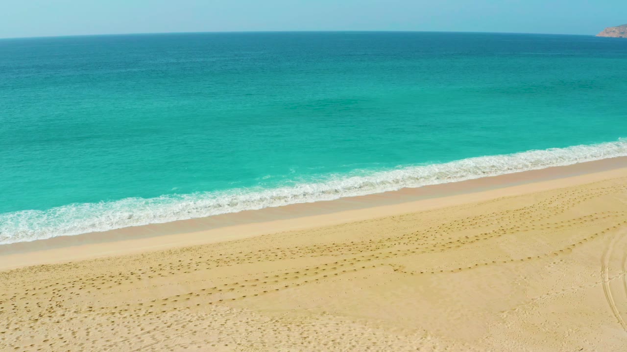Elevated Dreams Beach frame showing the surf line slightly farther along the sand, blue-green ocean bands, pale beach foreground, and no visible people.