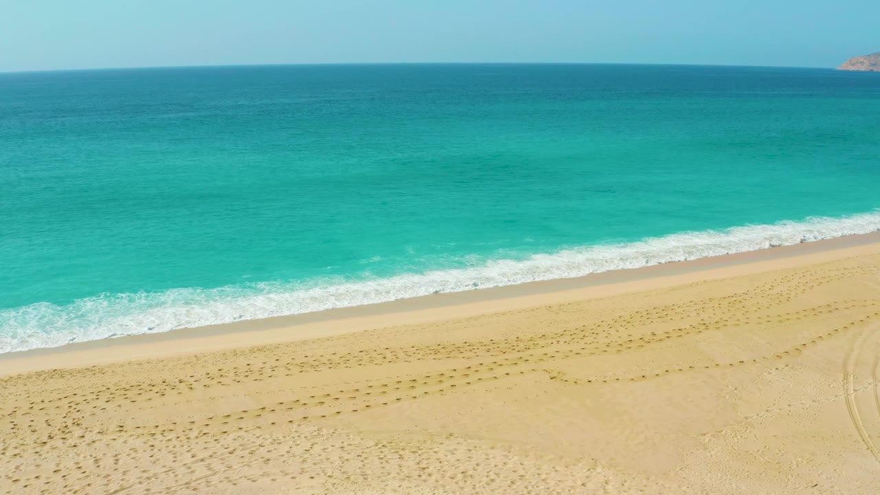 Aerial-looking view at Dreams Beach with more open sand in the foreground, a narrow white shore break, layered turquoise sea, and no visible people.