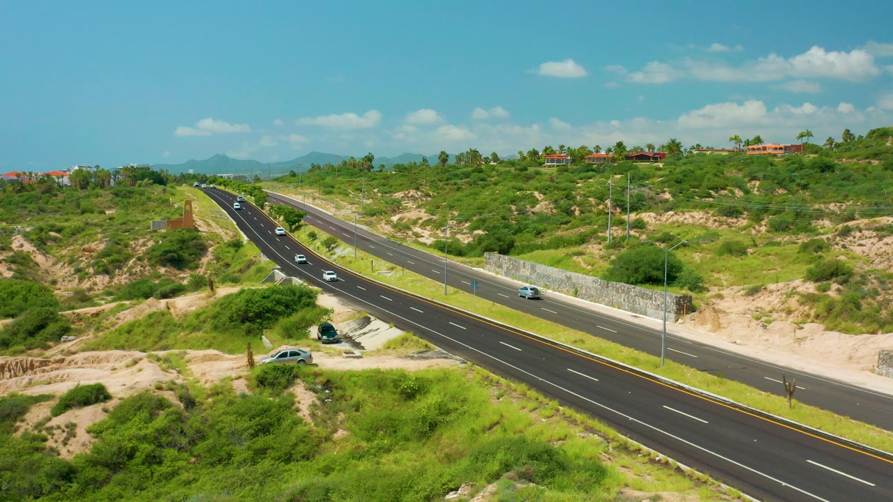 Aerial view over Dreams Beach, Baja California Sur, with cars traveling on a divided road through green scrub and sandy terrain, low hills in the distance, and no visible people.