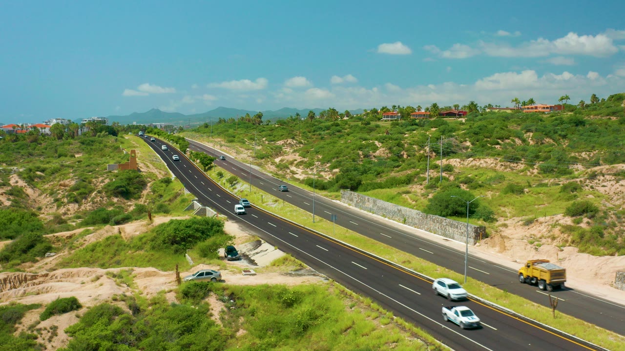 At Dreams Beach in Baja California Sur, Mexico, a divided black road cuts through green scrub and sandy ground with several cars and a yellow lorry visible, and no visible people.