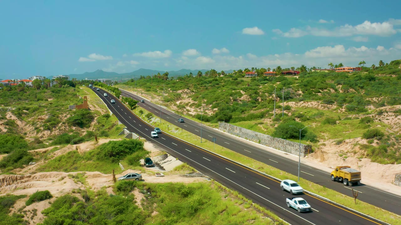 A high aerial frame at Dreams Beach shows cars spaced along the divided road, green brush on both sides, sandy embankments, distant hills, and no visible people.