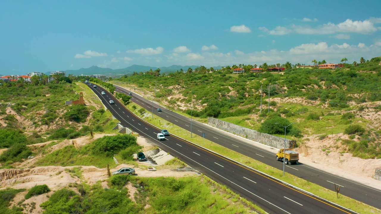 The Dreams Beach road remains framed from above as a yellow lorry advances on the right side, white cars travel the opposite lanes, and green scrubland fills the background with no visible people.