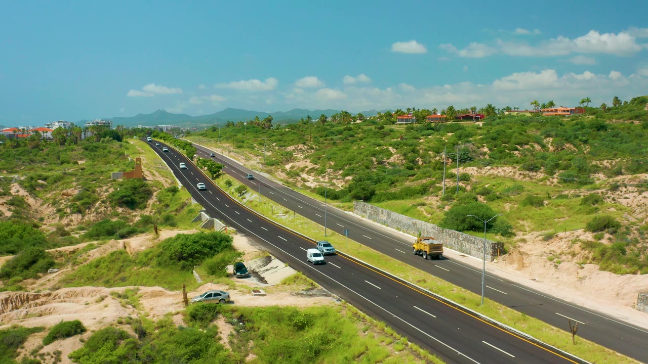Diagonal divided road with light traffic, a roadside wall, and green hills beyond.