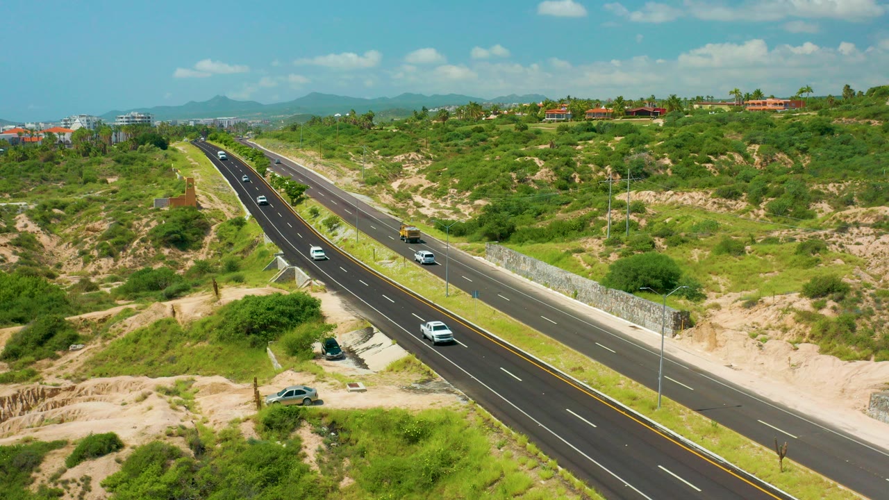 Opening frame of highway traffic in Dreams Beach, Baja California Sur, with cars on a divided road, sandy green hillsides, distant buildings, mountains, blue sky, and no visible people.