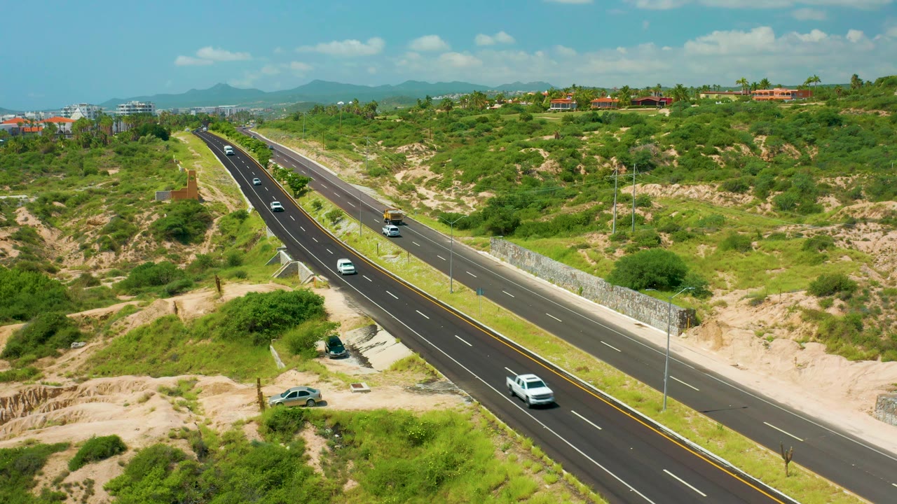 Aerial view over Dreams Beach, Baja California Sur, with a white car nearer the lower roadway, other vehicles farther up the divided highway, scrubby green terrain, distant hills, and no visible people.