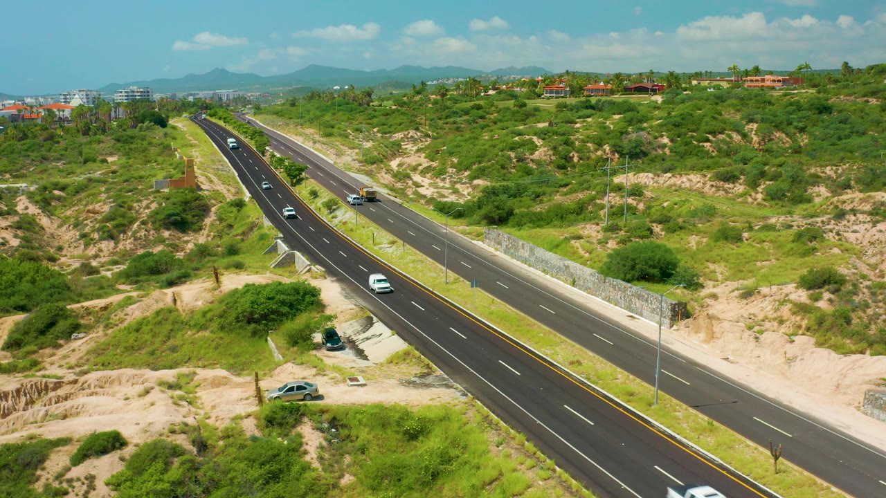 Divided highway curves through green and sandy hills with cars visible.