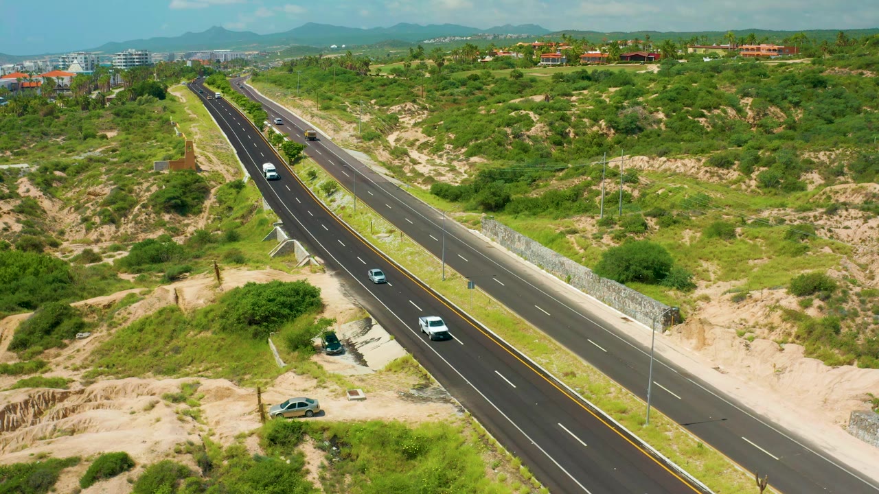 Elevated view of the Dreams Beach highway with a white vehicle and darker cars spaced along the lanes, scrubby green terrain on both sides, distant buildings, mountains, and no visible people.