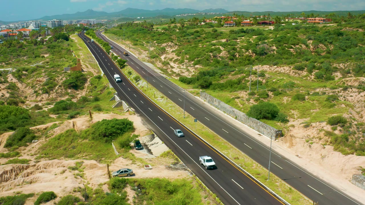 Aerial highway frame from Dreams Beach, Baja California Sur, showing a white van near the lower lanes, smaller cars farther up the road, green brush, sandy slopes, and no visible people.