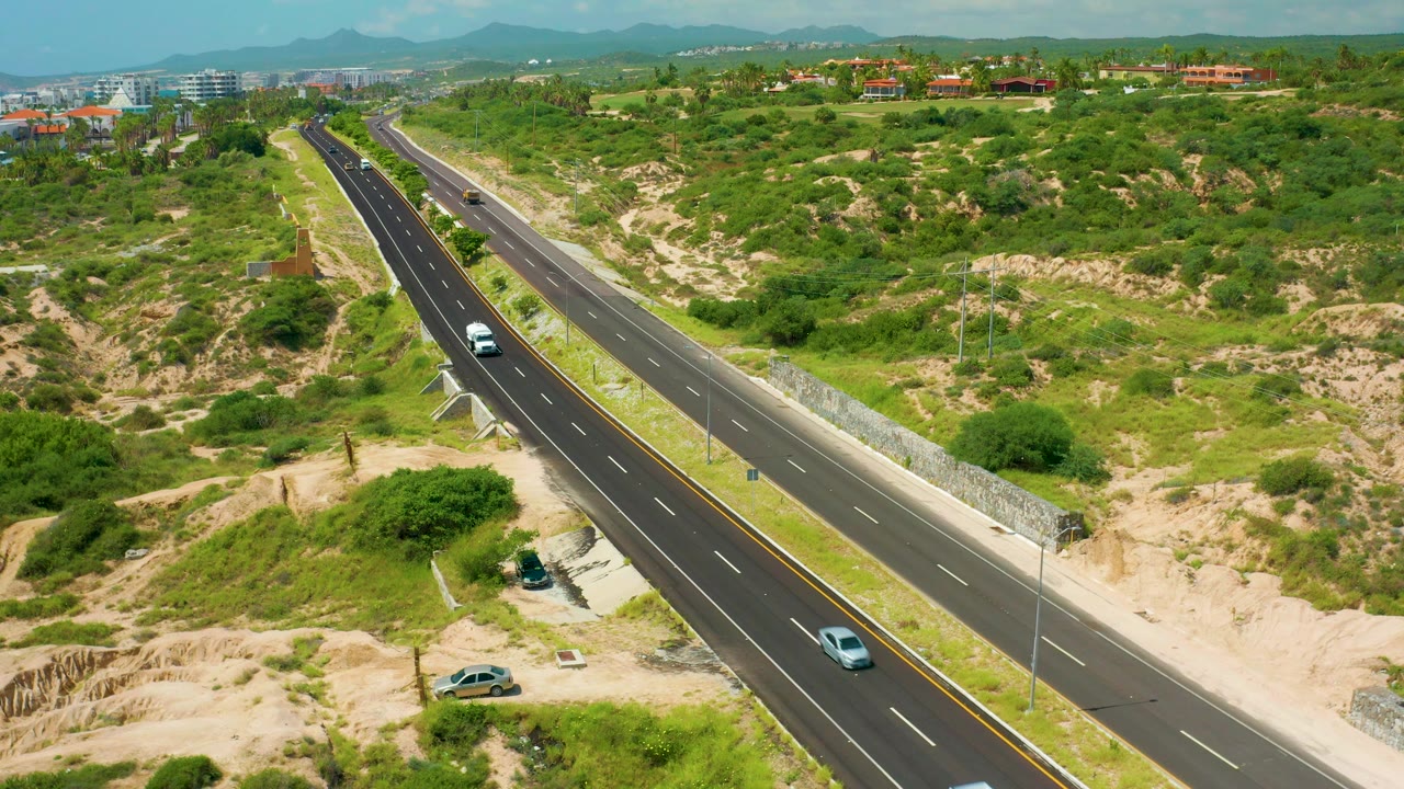 High-angle view of the highway at Dreams Beach with a white vehicle, a blue-gray car, dense green scrub, pale sandy ground, low distant buildings, mountains, and no visible people.
