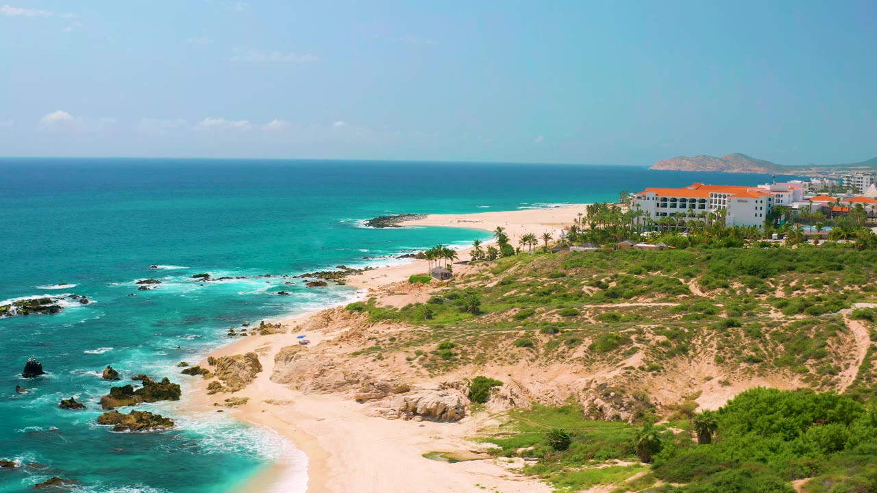 At 0:01, the aerial view keeps the Dreams Beach shoreline centered as white waves curl around dark rocks below the sandy point, with no visible people.