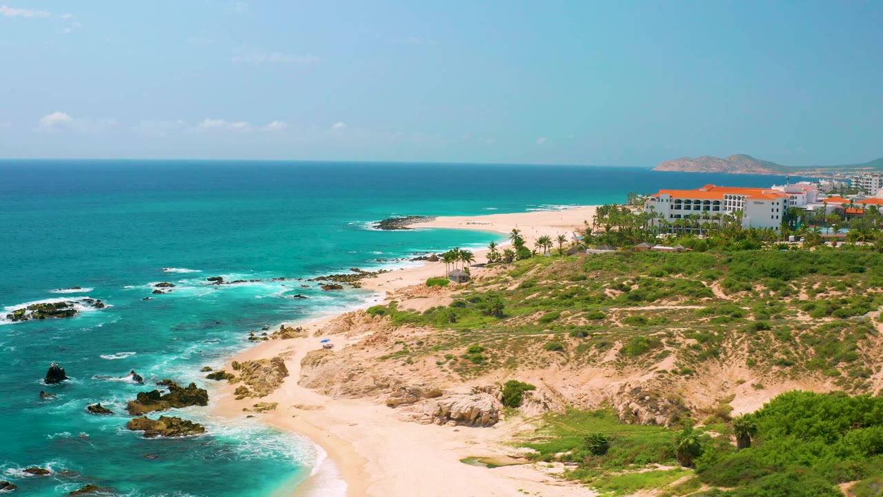 At 0:02, Dreams Beach fills the frame with the rocky promontory lower right, turquoise water left, a broad sand strip beyond, and no visible people.