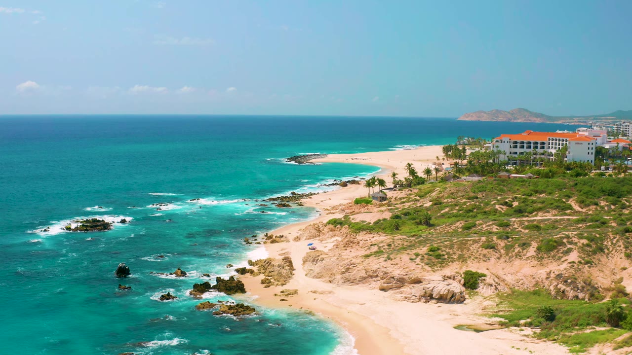At 0:01, the Dreams Beach aerial angle holds on surf breaking around dark rocks, a curving white beach, tan bluff, resort buildings, and no visible people.