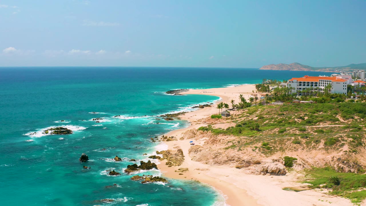 At 0:03, a wide aerial frame over Dreams Beach shows foamy water around scattered rocks, white sand, dry scrubland, resort buildings, and no visible people.