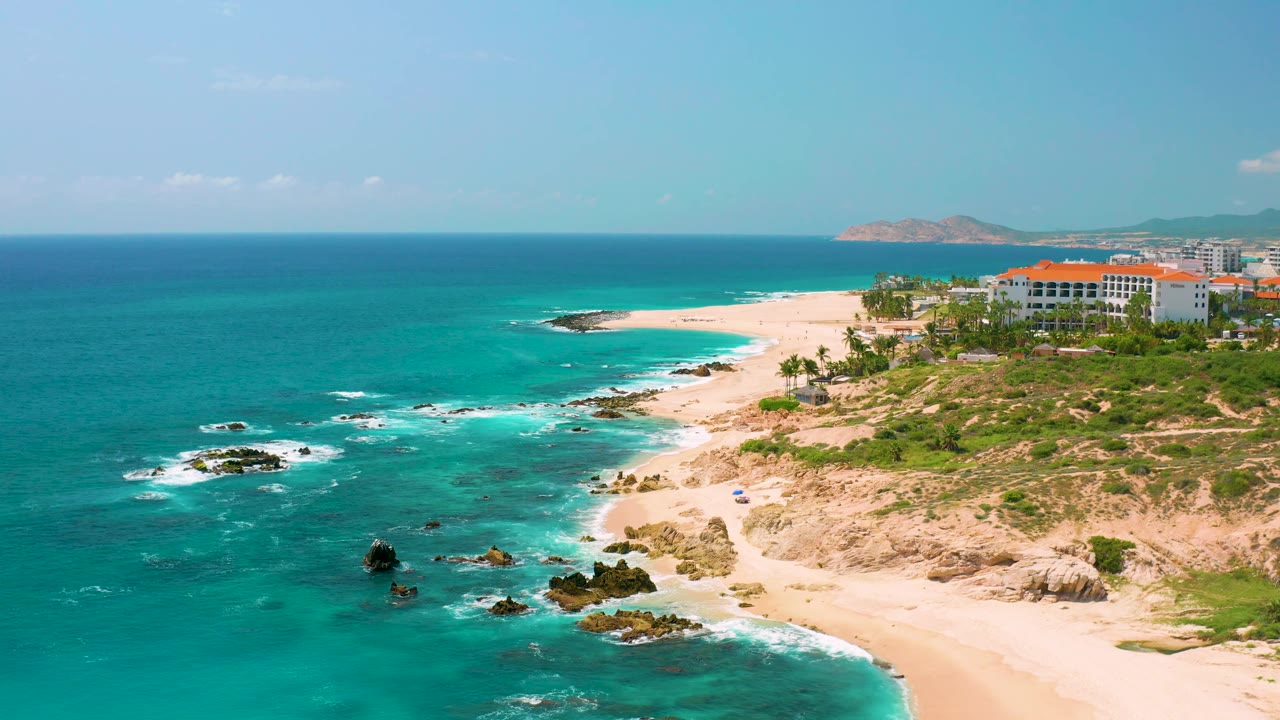 Dreams Beach coastline in Baja California Sur with open turquoise water, rocky surf breaks in the lower frame, white beach sweeping toward buildings, and no visible people.
