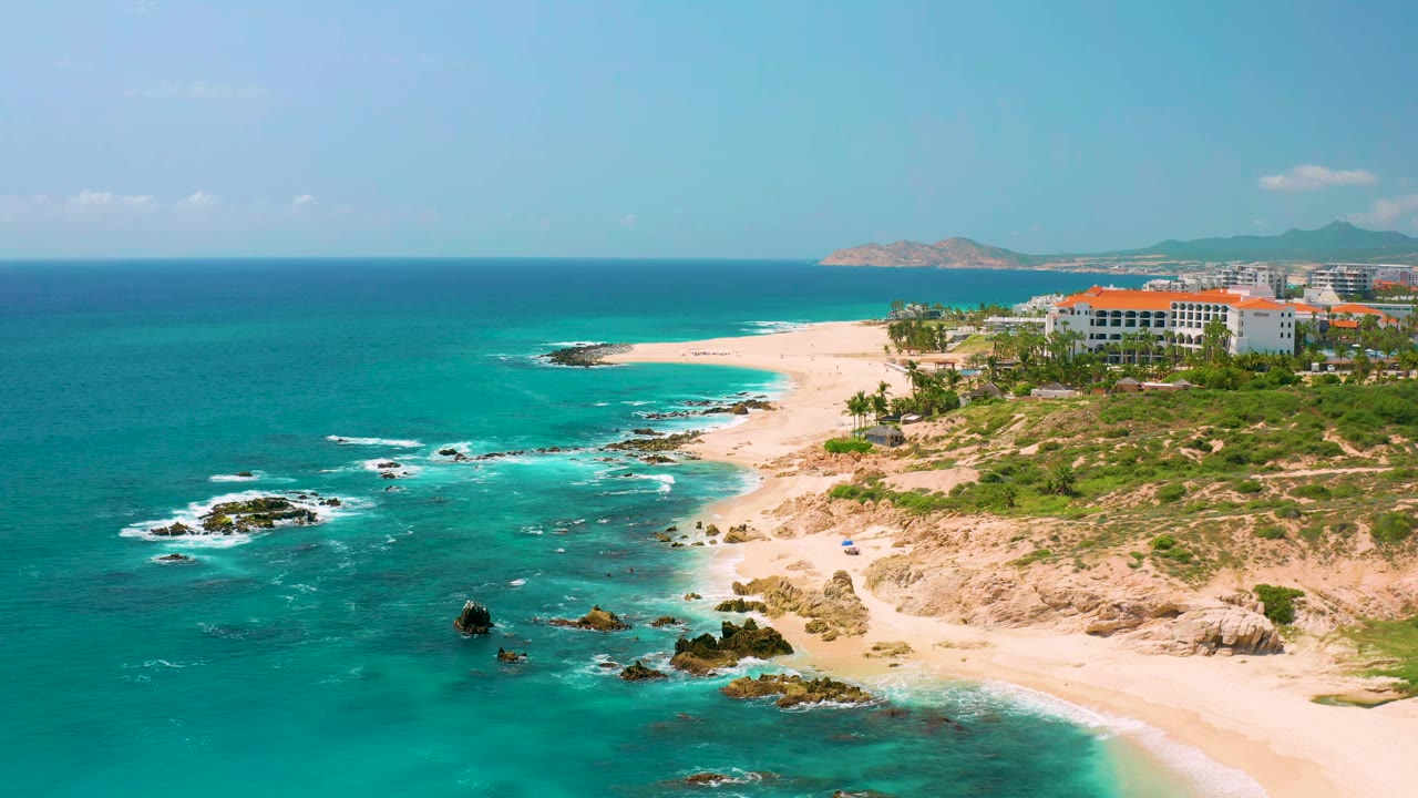 Aerial frame over Dreams Beach, Baja California Sur, Mexico, showing white surf lines pushing toward sandy shoreline, scattered rocks in clear water, resort-style buildings to the right, and no visible people.