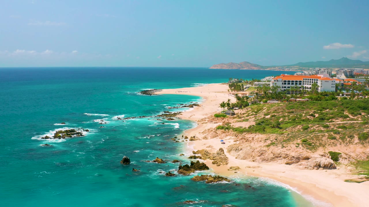 Aerial view of Dreams Beach in Baja California Sur, Mexico, with deeper blue water near the horizon, bright turquoise shallows, pale beach, rocky outcrops, green scrub, buildings at right, and no visible people.