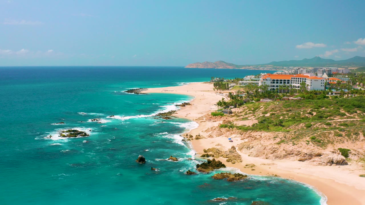 Opening aerial frame over Dreams Beach, Baja California Sur, with turquoise water, scattered rocks, an empty sand beach, coastal buildings, distant mountains, and no visible people.