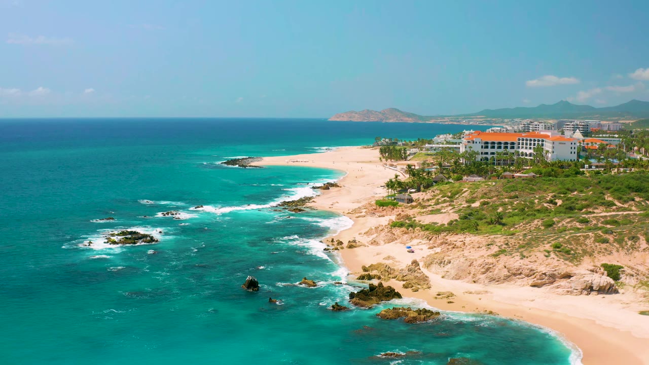 One second into the aerial view, Dreams Beach shows clear blue water in the foreground, surf breaking around rocks, pale sand, large white coastal buildings, mountains, and no visible people.
