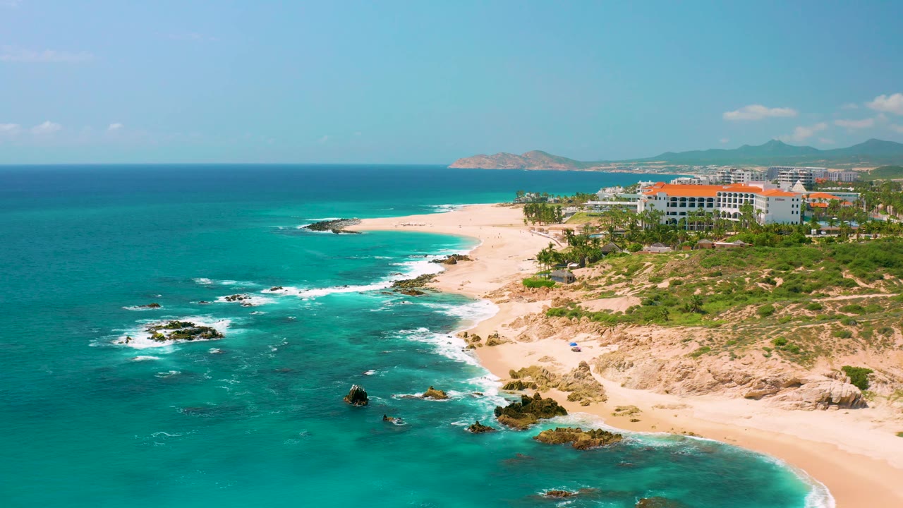 At 0:02, the frame emphasizes the turquoise sea beside an empty Dreams Beach shoreline, with dark rocks, white wave edges, coastal greenery, buildings, mountains, and no visible people.