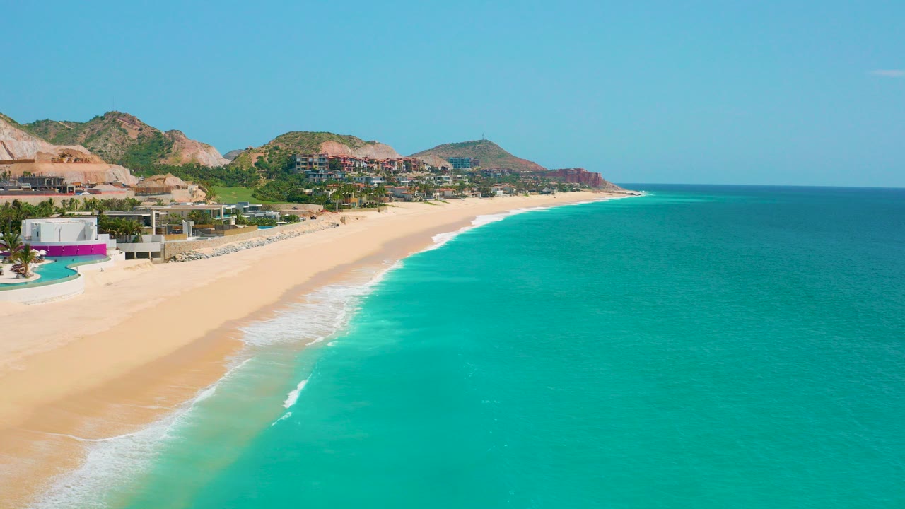 Opening frame over Dreams Beach in Baja California Sur, Mexico, showing turquoise water, pale sand, hillside homes, and no visible people.