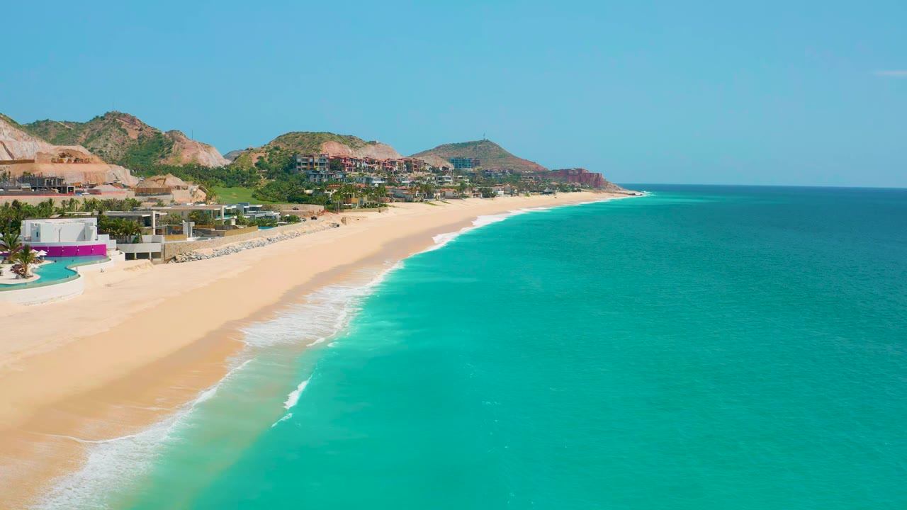 Drone frame at Dreams Beach in Baja California Sur, Mexico, with the surf line cutting diagonally past sand, coastal houses, green hills, and no visible people.