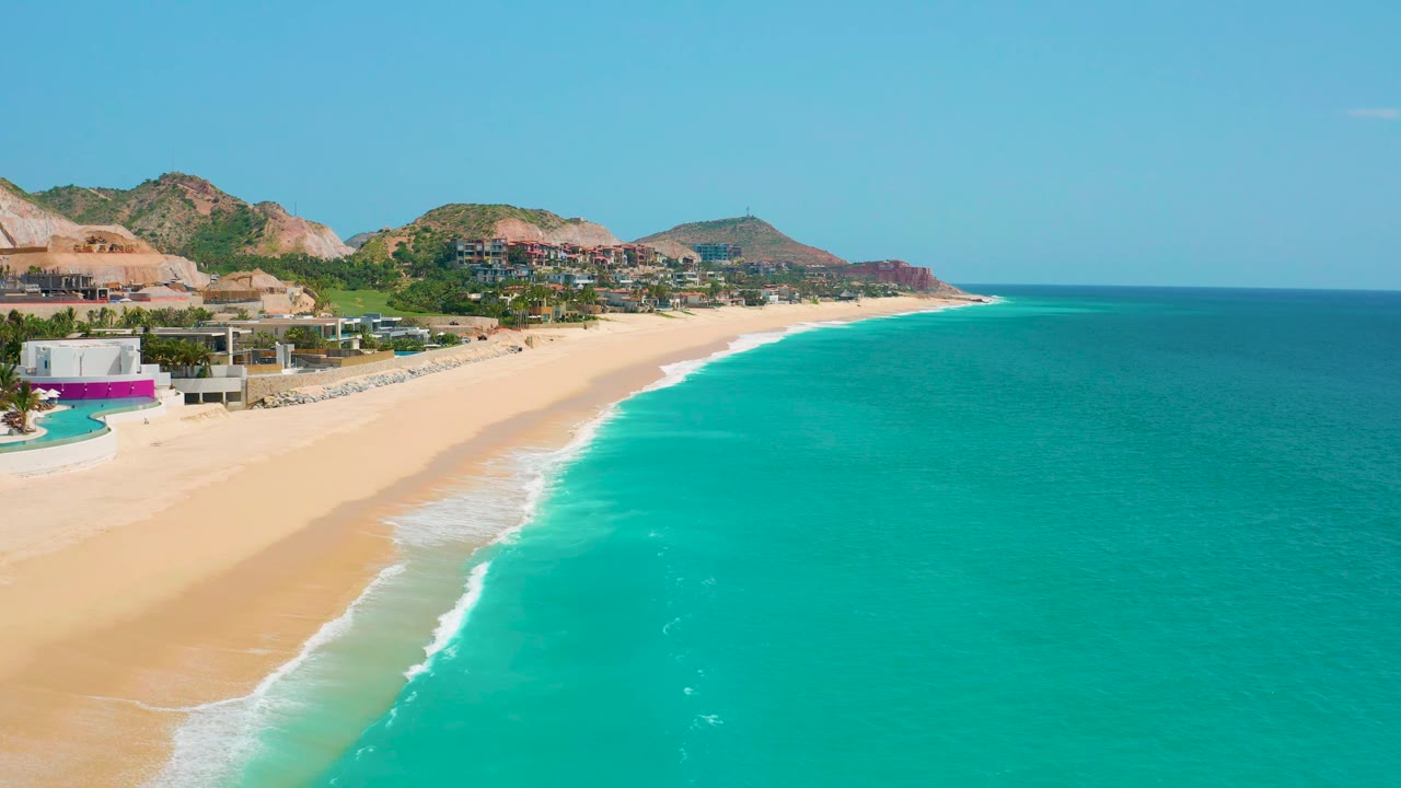 Elevated frame of Dreams Beach in Baja California Sur, Mexico, with more open water on the right, the beach and hillside buildings to the left, and no visible people.