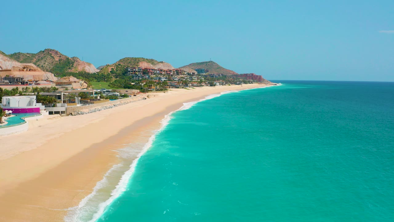 Aerial coastline frame with beach residences, sand, surf, and blue-green water.