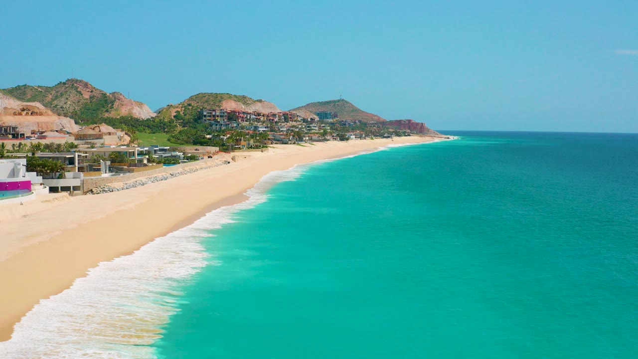 Opening aerial frame over Dreams Beach in Baja California Sur, with turquoise sea on the right, pale sand and white surf on the left, hillside buildings behind the shore, and no visible people.