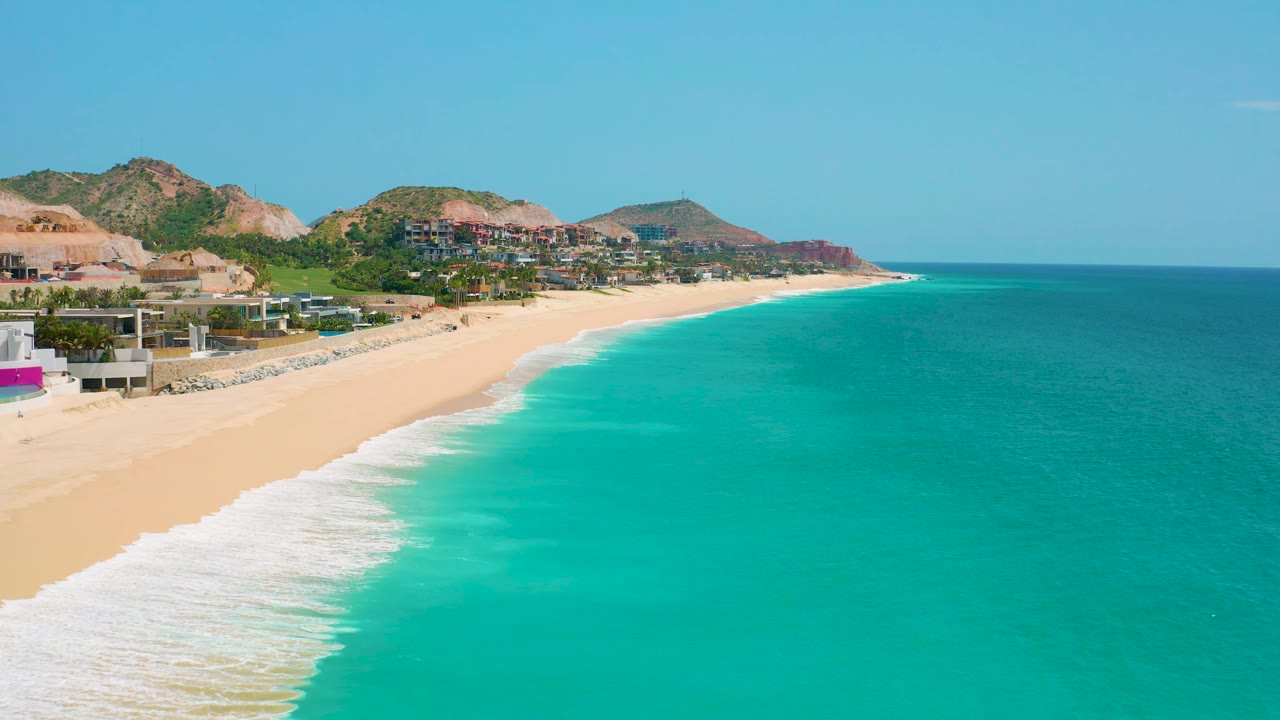 Aerial view of Dreams Beach with the coastline receding toward the background, bright turquoise water filling the lower right, pale sand along the left, hillside buildings beyond the shore, and no visible people.