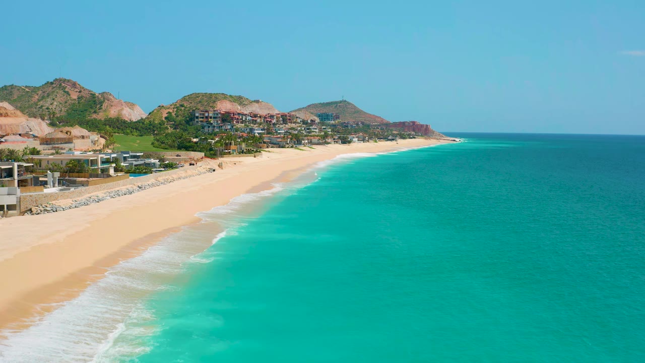 Dreams Beach shoreline curves through the frame with pale sand, bright cyan water, hillside greenery, clustered buildings, and no visible people.