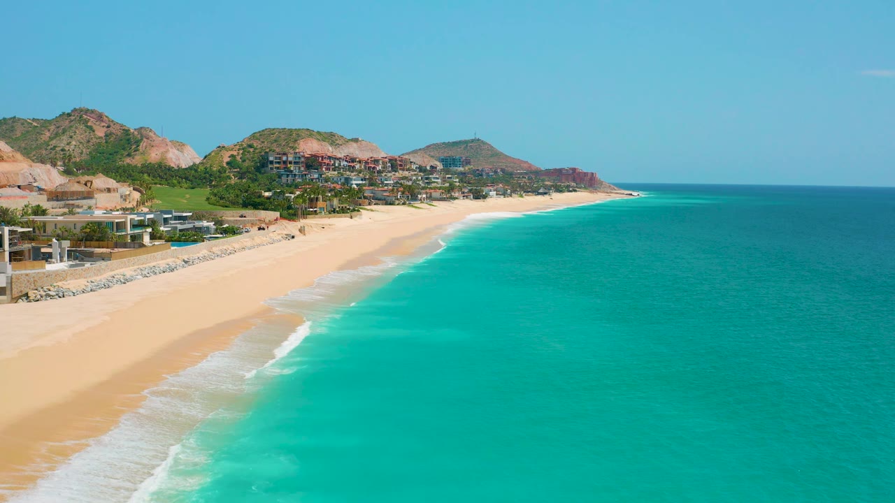 A wide Dreams Beach view shows more open turquoise water to the right, a narrow white surf line, sand at left, hillside buildings, and no visible people.