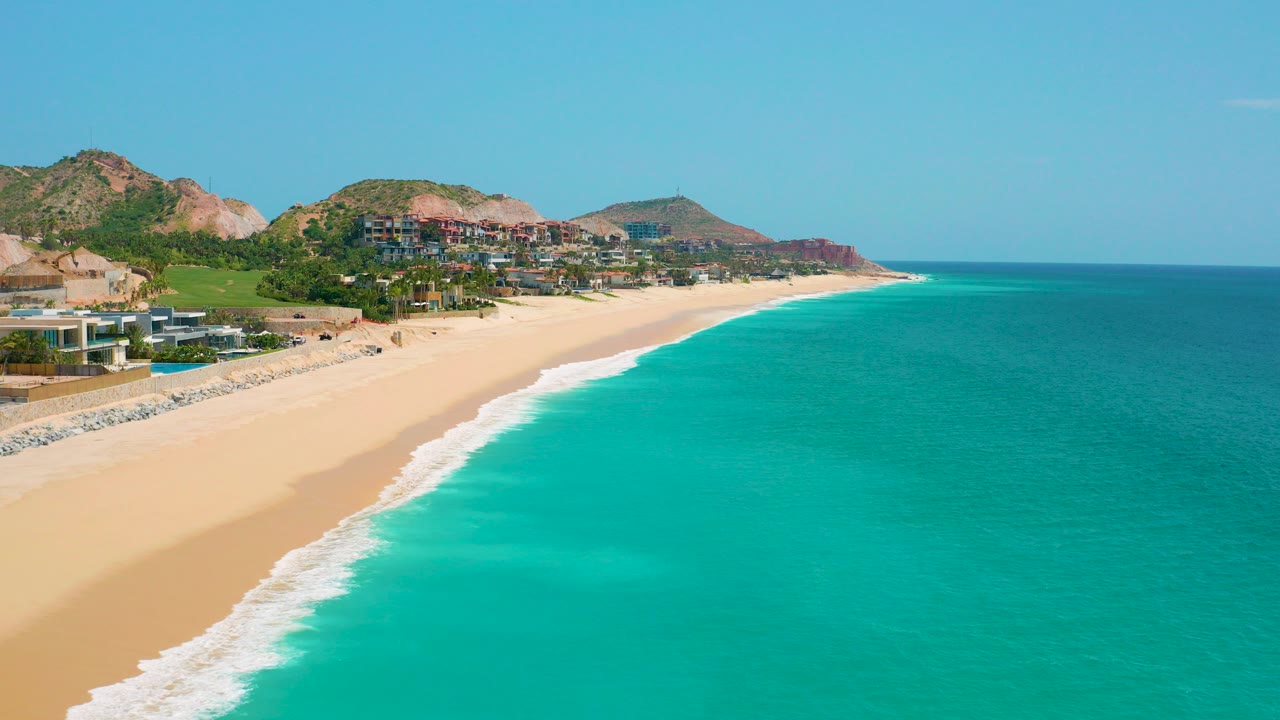 Opening aerial frame of Dreams Beach in Baja California Sur, Mexico, with pale sand at left, turquoise ocean at right, hillside buildings beyond the shore, and no visible people.