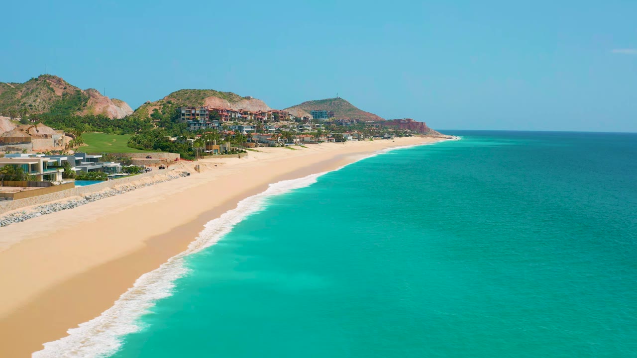 Wide beach shoreline with white surf and turquoise water beside hillside development.