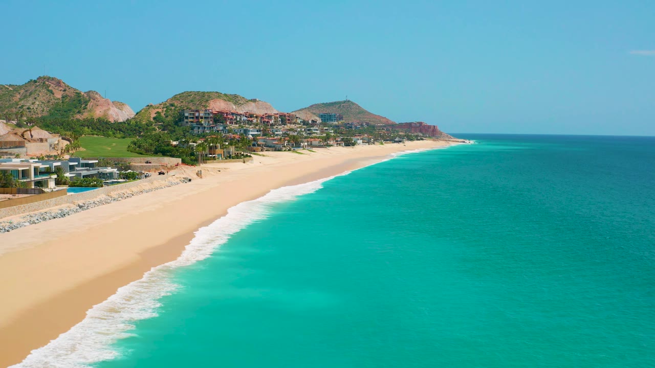 At 0:02, the aerial frame keeps the ocean dominant on the right while the sand, surf line, hillside structures, and rocky slopes remain visible at Dreams Beach, with no visible people.