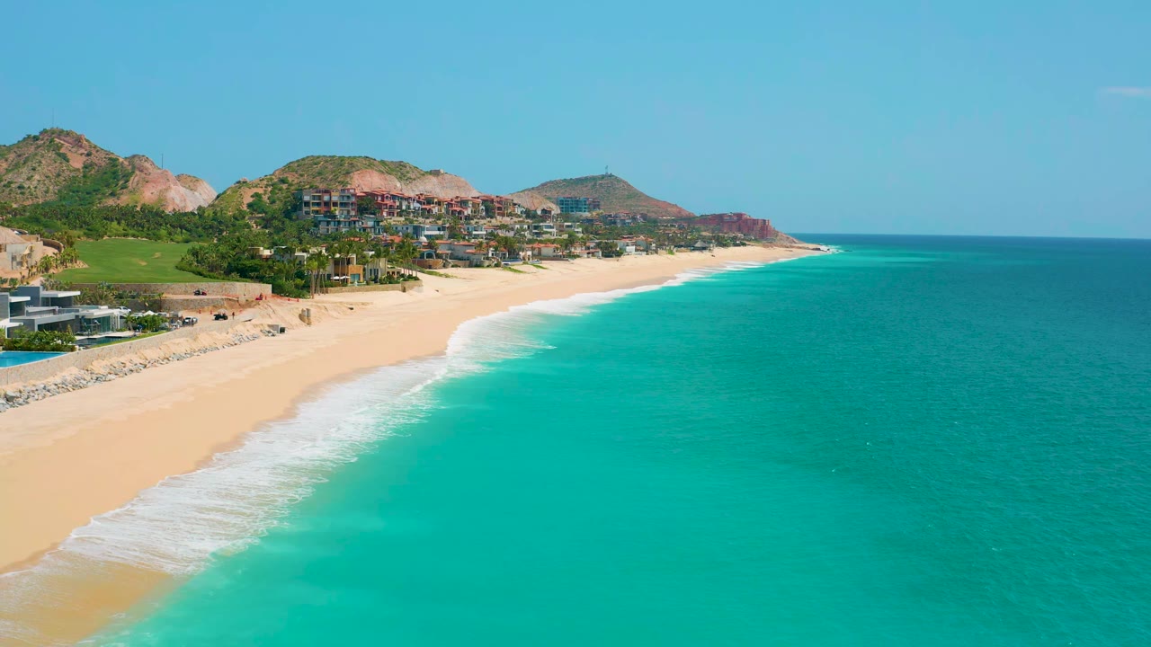 Wide shoreline with sandy beach, rolling surf, turquoise sea, and hillside buildings.