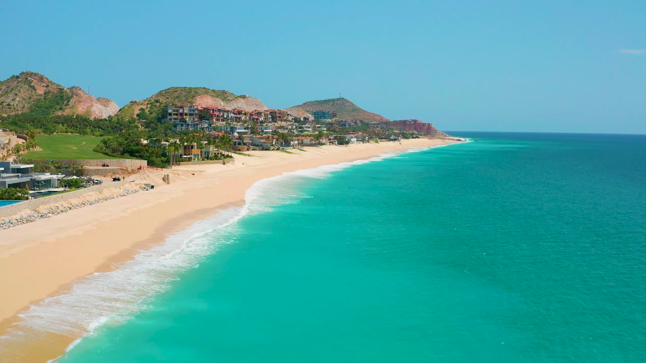 Aerial view of Dreams Beach in Baja California Sur, Mexico, where a wider stretch of turquoise water fills the right side, the pale beach narrows toward the distance, and no visible people appear on the sand.