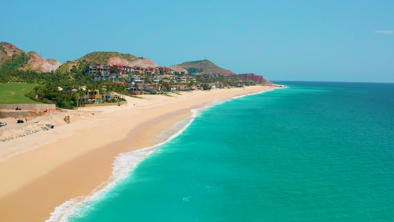 Opening frame over Dreams Beach in Baja California Sur, Mexico, with turquoise water on the right, a pale sandy shoreline on the left, green hills and clustered buildings in the background, and no visible people.
