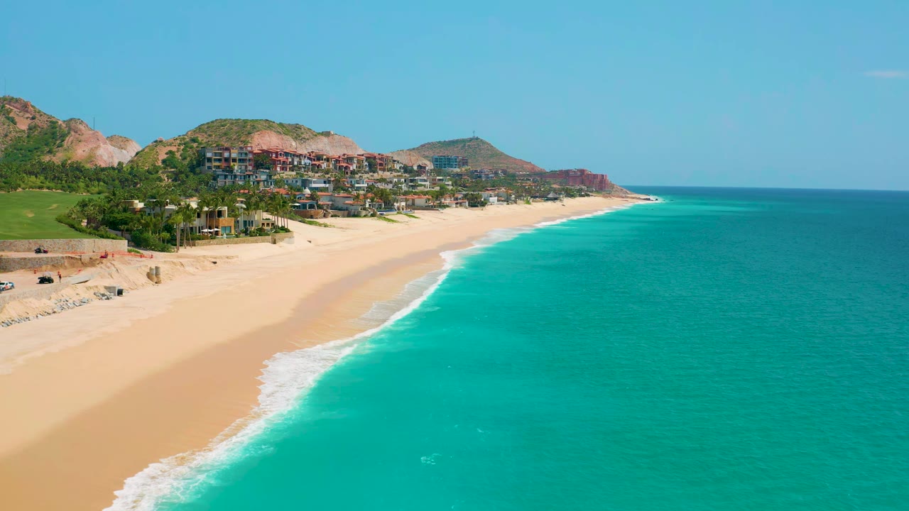 At 0:01, the Dreams Beach shoreline fills more of the center, with a white wave line tracing the sand, turquoise sea to the right, hillside buildings above the beach, and no visible people.
