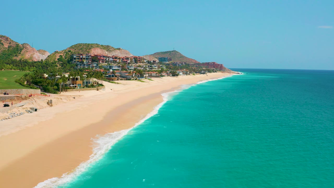 At 0:02, the beach and turquoise ocean remain in a broad aerial composition, with the curving coast leading toward a distant headland and hillside buildings visible above the sand, with no visible people.