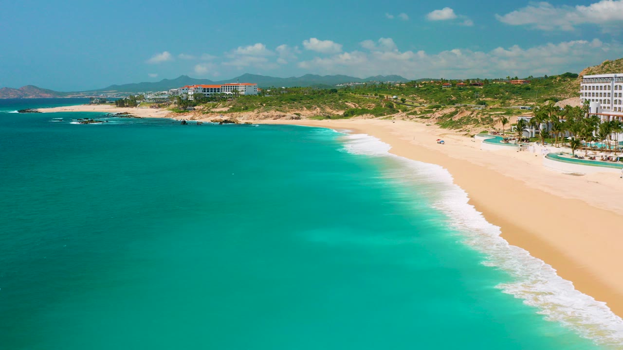 Wide aerial view of Dreams Beach in Baja California Sur, Mexico, showing turquoise water, pale sand, surf, green dunes, distant resort buildings, and no visible people.
