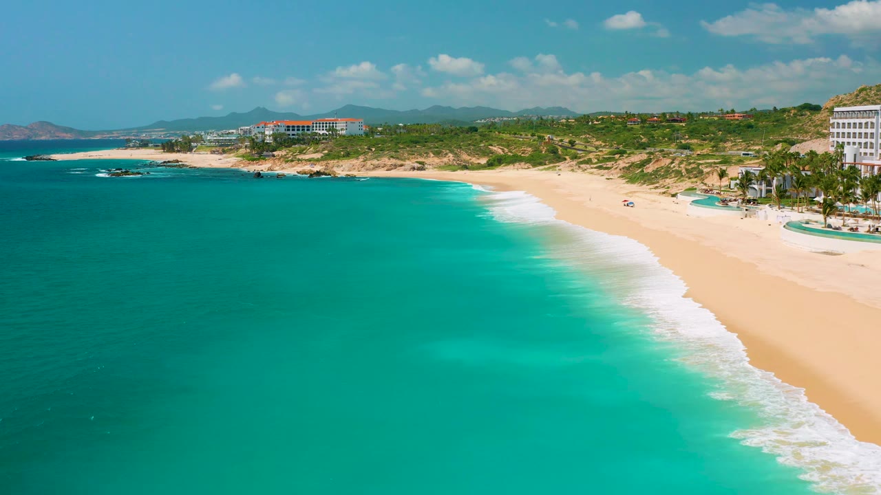 Dreams Beach aerial view in Baja California Sur, Mexico, showing smooth turquoise water, a diagonal band of white surf, pale sand, low greenery, a resort edge, and no visible people.