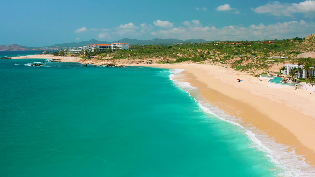 Aerial view at Dreams Beach in Baja California Sur, Mexico, with turquoise sea on the left, a pale sand beach curving right, green vegetation inland, distant hills, and no visible people.