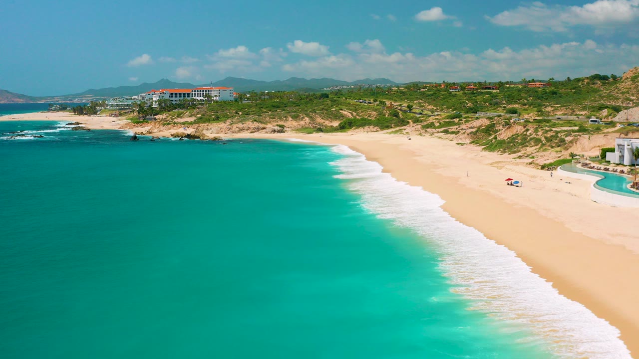 Opening aerial frame over Dreams Beach in Baja California Sur, Mexico, with turquoise water in the foreground, white surf along the sand, resort buildings beyond the beach, and tiny people visible near umbrellas.