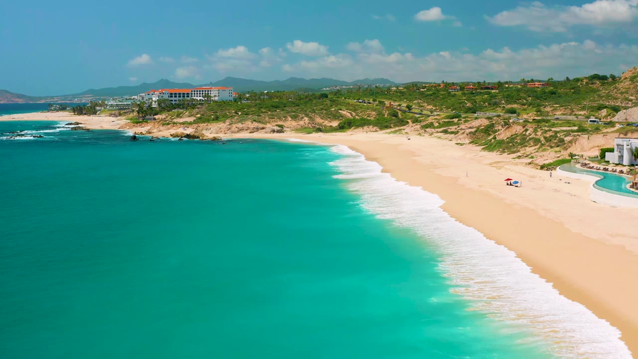 Wide drone view of a curved beach with surf, turquoise water, and hillside resort area.