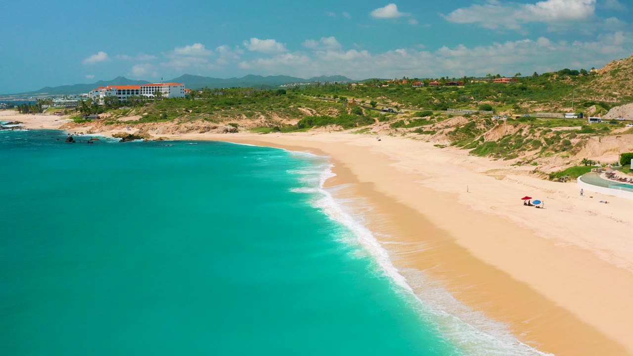 Aerial frame over Dreams Beach in Baja California Sur with turquoise water at left, a pale sand shoreline at right, low surf, dunes, a large coastal building, distant hills, and no visible people.