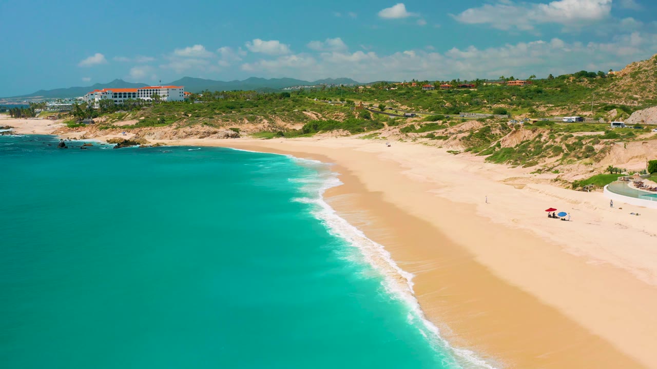 Aerial frame over Dreams Beach with blue-green water filling more of the left side, a narrow white wave edge on the sand, dunes and roads inland, a coastal building near the left horizon, and no visible people.