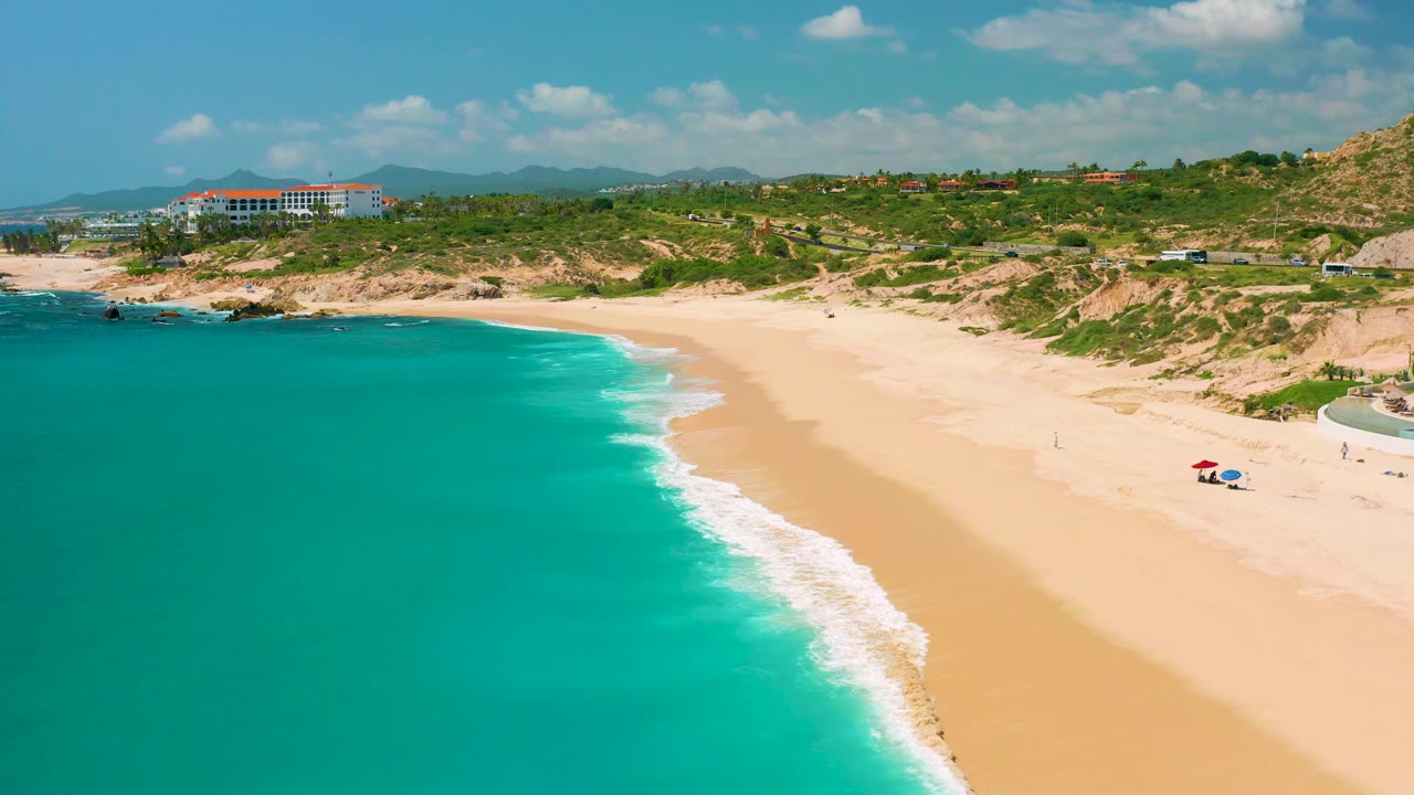 Aerial shoreline frame with mild waves, open sand, dunes, and distant hills.