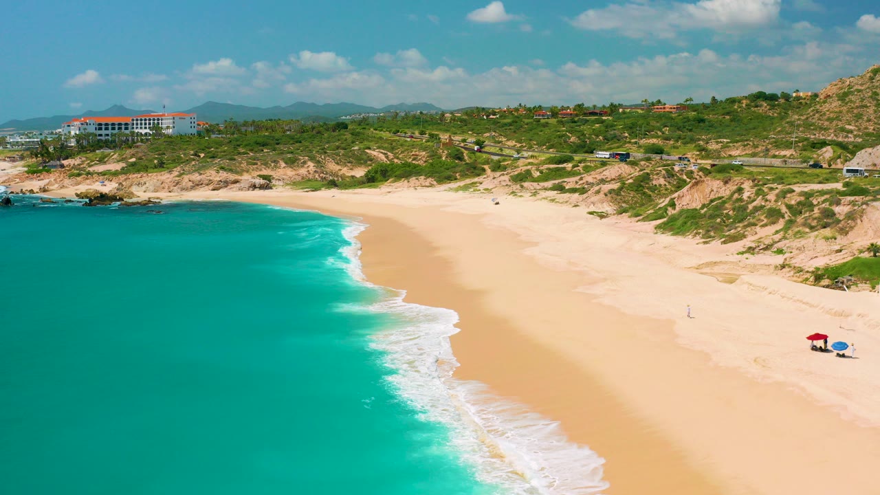 Aerial frame over Dreams Beach in Baja California Sur, with turquoise water at left, a pale surf edge, yellow sand, scrubby dunes, distant buildings, and no recognizable people.